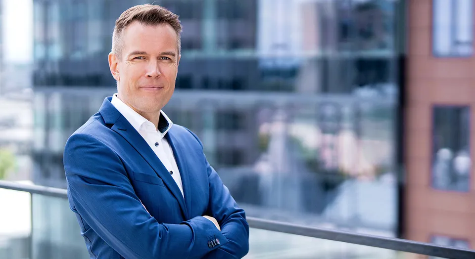Ein Mann mit braunen Haaren den man Oberkörper aufwärts abgebildet sieht, trägt einen dunkelblauen Anzug mit dunkler Krawatte und lächelt in die Kamera