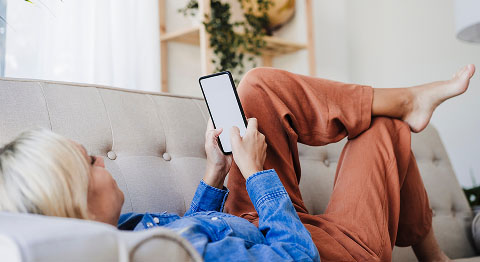 Blonde Frau in Jeanshemd liegt auf beigem Sofa und schaut in ihr Smartphone