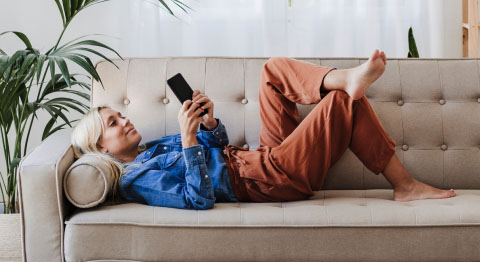 Blonde Frau in Jeansjacke liegt auf beigem Sofa und schaut in ihr Smartphone