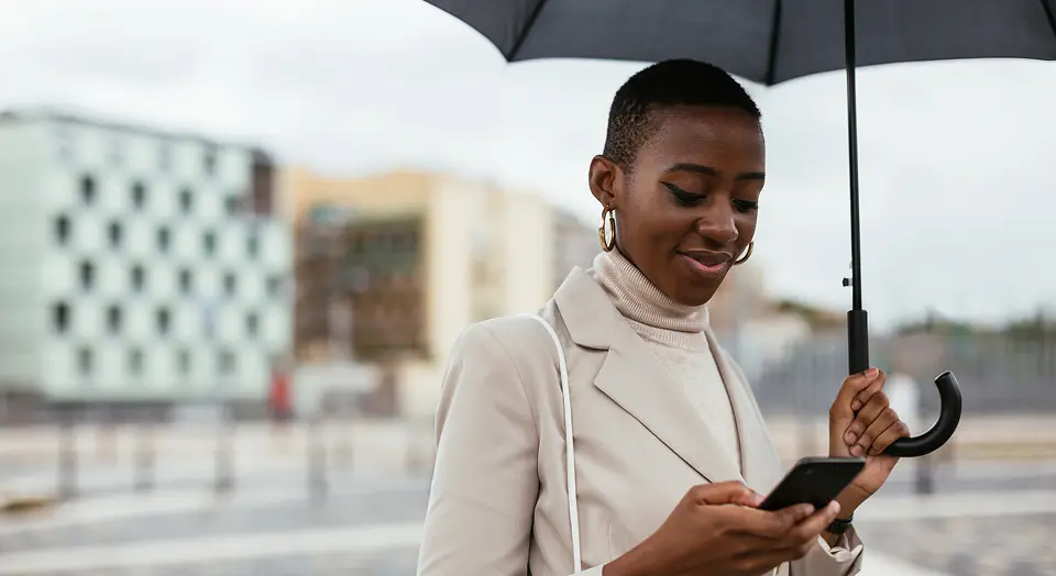 Frau mit Mantel und Regenschirm steht im Freien und schaut auf ihr Smartphone