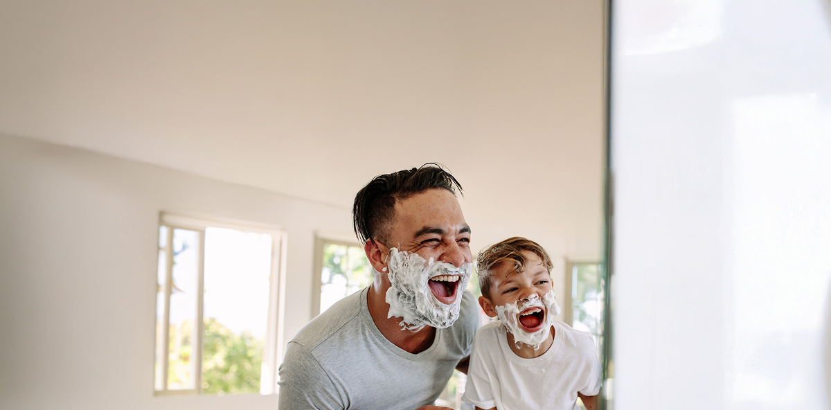 Vater und Sohn mit Rasierschaum im Gesicht schauen in einen Spiegel und lachen